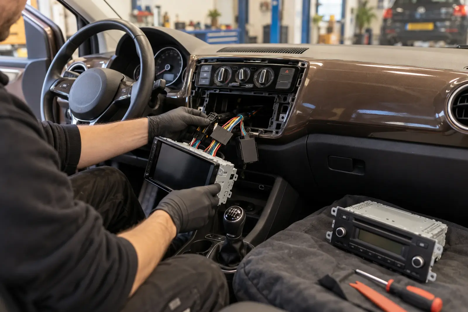 Android Auto radio inbouwen in Venlo: monteur met handschoenen houdt de nieuwe 2-DIN autoradio met kabelboom voor de open radio-opening van een Volkswagen up, de oude radio ligt op de stoel